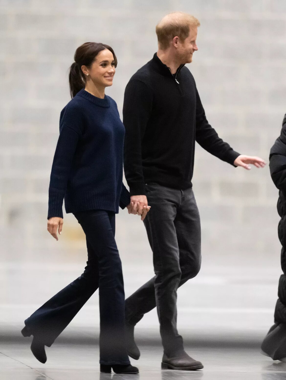 Meghan Markle wears a navy sweater, black bootcut jeans, and black boots while walking with Prince Harry